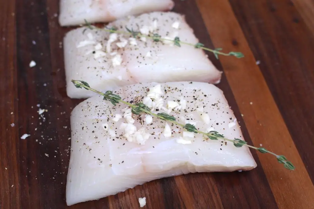 Filetti di halibut freschi conditi con aglio, timo, sale e pepe su un tagliere di legno.