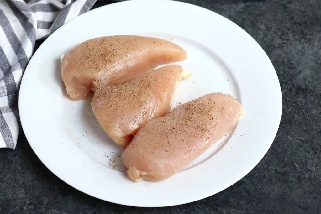 Petti di pollo crudo conditi con sale, pepe e aglio in polvere su un piatto bianco.